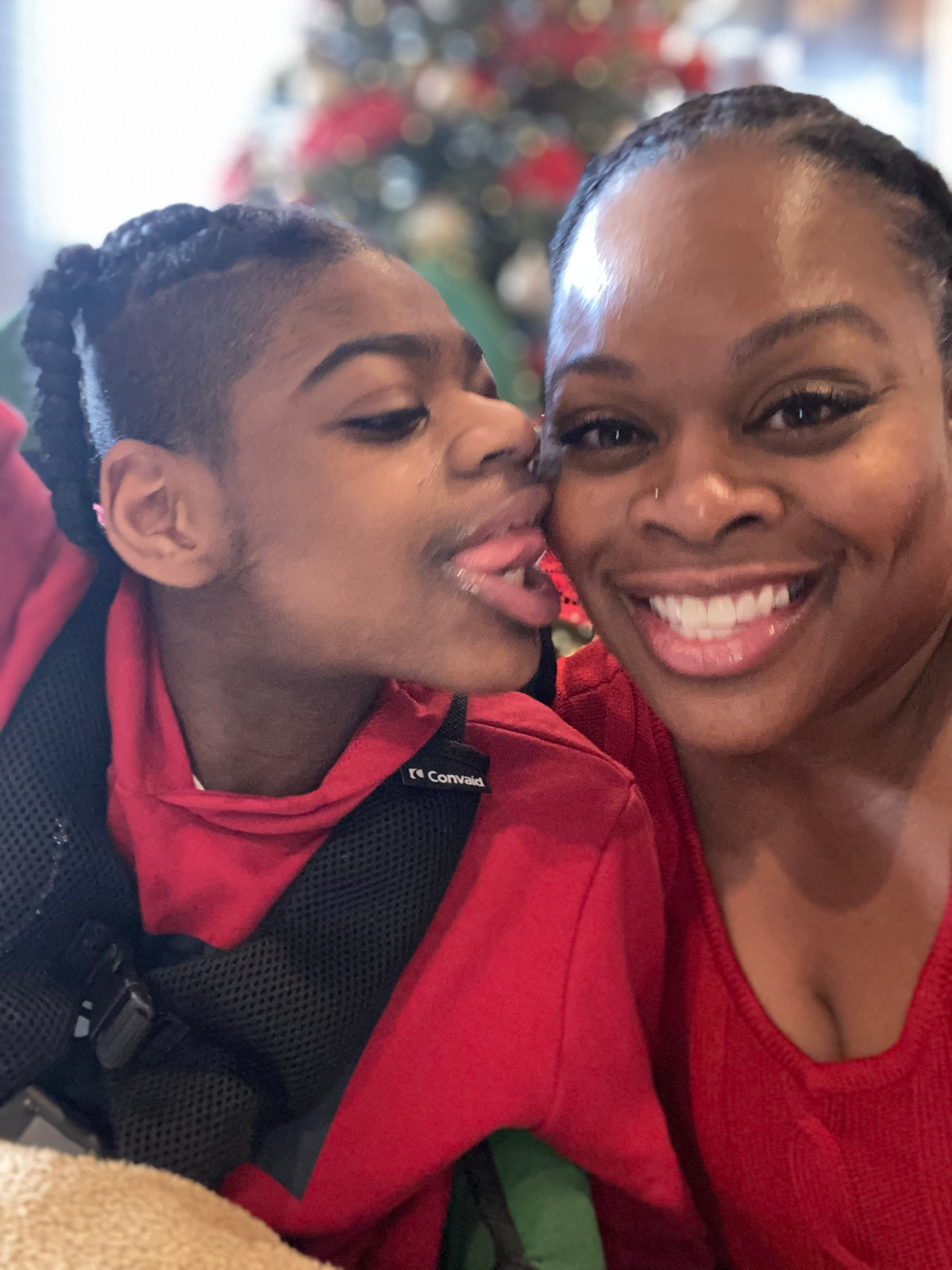 Young boy in wheelchair kissing his mother on the cheek in front of the Chrismas tree.
